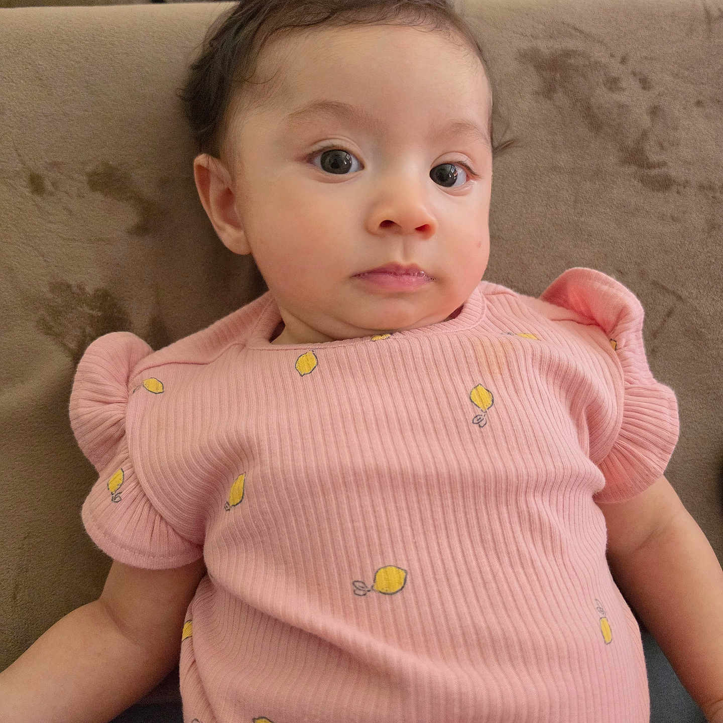 Amyra is registered to the contest to win money with this photo: baby, child, person, infant, pink_clothing, lemon_pattern, sitting, couch, furniture, indoor, portrait, face, curly_hair, expression, young_child, cute, skin, head, eyes, arm