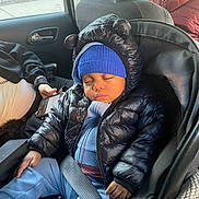 Kayden joined the competition — help win amazing prizes! baby, child, sleeping, car_seat, jacket, hat, blue_clothing, puffer_jacket, seatbelt, car_interior, person, hand, phone, window, road, fence, seat, cozy, cute, toddler