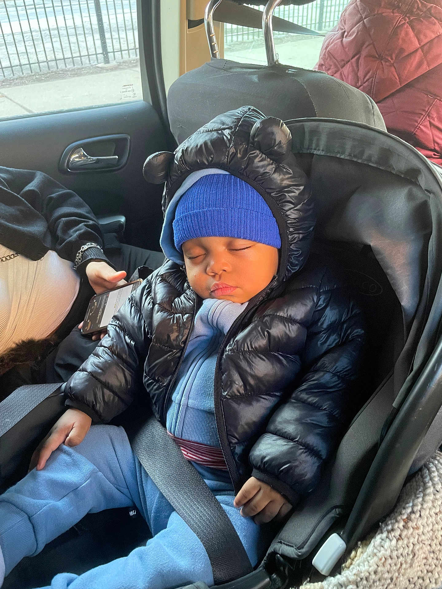 Kayden joined the competition — help win amazing prizes! baby, child, sleeping, car_seat, jacket, hat, blue_clothing, puffer_jacket, seatbelt, car_interior, person, hand, phone, window, road, fence, seat, cozy, cute, toddler