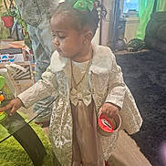 Amria joined the competition — help win amazing prizes! carpet, child, coat, curious, curly_hair, decor, dress, footwear, furniture, glass_table, green_bow, home, indoor, jewelry, living_room, person, standing, toddler, toy, vacuum_cleaner