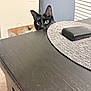 cat, black_cat, pet, peeking, dining_table, placemat, chair, wood_table, green_eyes, ears, whiskers, indoor, curious, close_up, home, furniture, tabletop_object, domestic_animal, portrait, sneaky