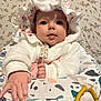 baby, infant, bonnet, hood, white_jacket, big_eyes, rosy_cheeks, hand, bouncer_seat, toy_rings, floral_background, patterned_quilt, soft_texture, sitting, portrait, indoor, cute, curious_expression, gaze, clothing