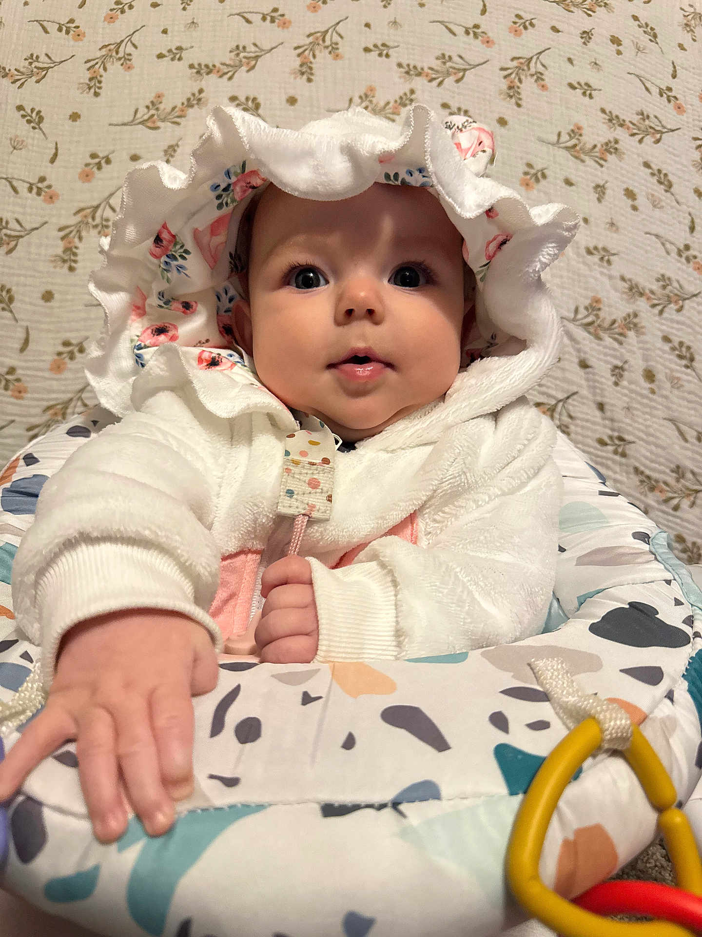 Leah joined the competition — help win amazing prizes! baby, infant, bonnet, hood, white_jacket, big_eyes, rosy_cheeks, hand, bouncer_seat, toy_rings, floral_background, patterned_quilt, soft_texture, sitting, portrait, indoor, cute, curious_expression, gaze, clothing