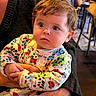 baby, child, infant, person, pajamas, colorful_clothing, curly_hair, indoor, hand, adult, table, wood, face, expression, seated, looking_away, cute, warm_lighting, human, holding