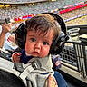 Tyler is registered to the contest to win money with this photo: baby, child, headphones, carrier, stadium, crowd, hand, person, seat, blurred_background, clothing, noise_reduction, curious, indoor, event, spectator, young_child, face, closeup, support
