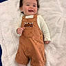 baby, child, smiling, curly_hair, orange_overalls, monster_print, white_towel, lying_down, cute, happy, infant, clothing, soft_texture, portrait, indoors, small_hands, baby_fashion, playful, cheerful, person