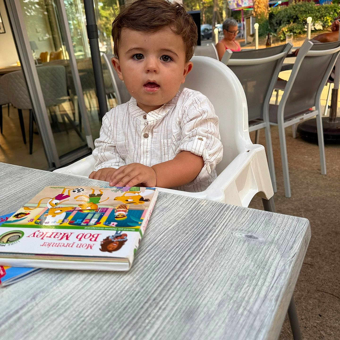 Raphaël participe au concours pour gagner de l'argent avec cette photo : background_people, book, brown_hair, cafe, casual_clothing, child, curious, daylight, glasses, high_chair, outdoor, person, reading, relaxing, seated, short_sleeves, sunlight, table, toddler, trees