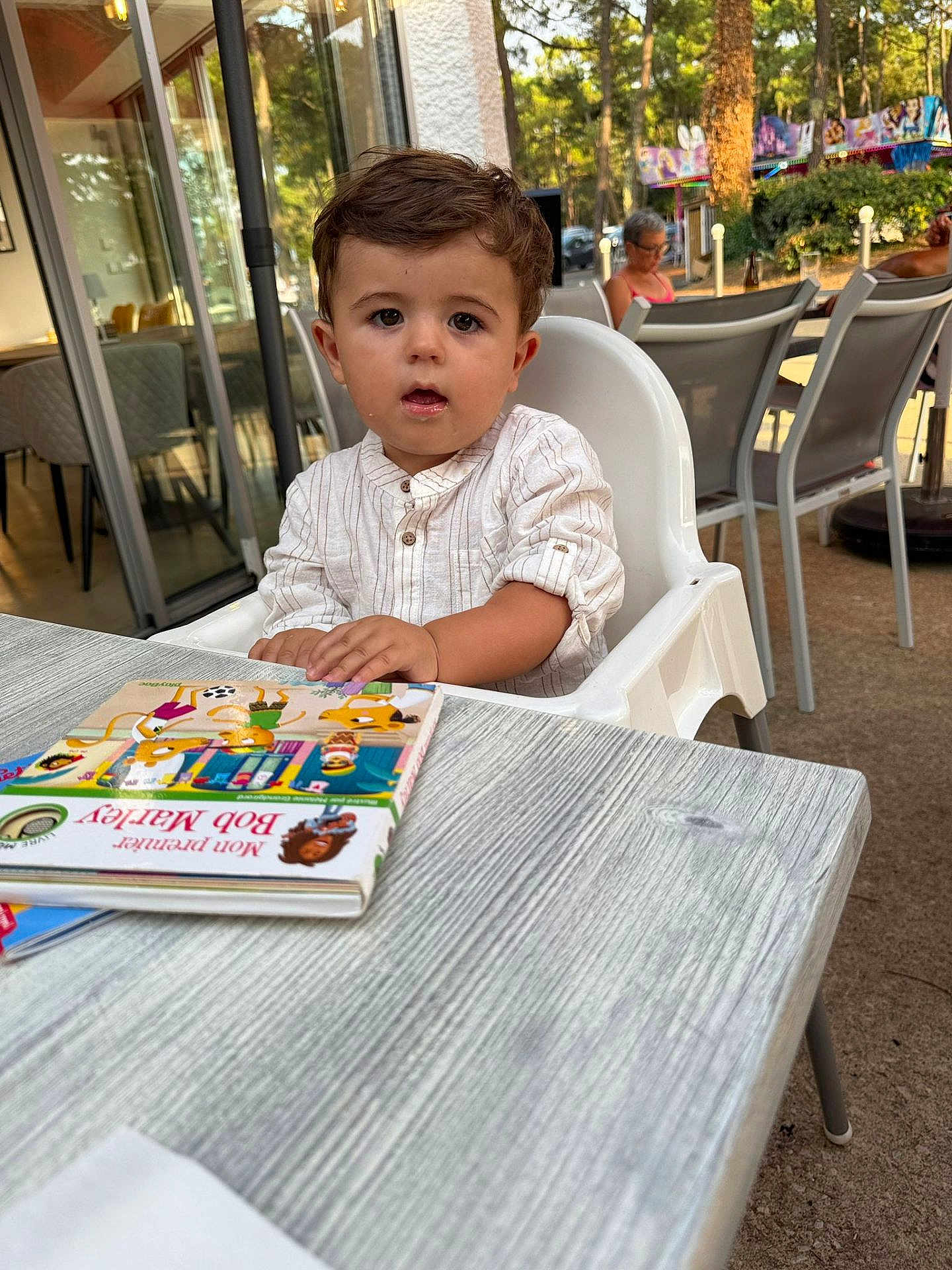Raphaël participe au concours pour gagner de l'argent avec cette photo : toddler, child, high_chair, book, table, outdoor, cafe, person, seated, curious, brown_hair, short_sleeves, sunlight, trees, background_people, glasses, relaxing, daylight, reading, casual_clothing