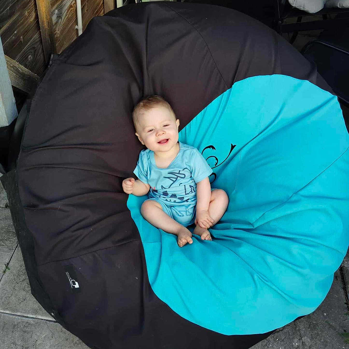 Gabriel a rejoint le concours — aidez-le/la à gagner de superbes lots ! baby, bean_bag, blue_clothing, casual, chair, child, comfort, floor, furniture, happy, infant, outdoor, person, relaxed, seat, sitting, smiling, stone_tiles, sunlight, wooden_wall