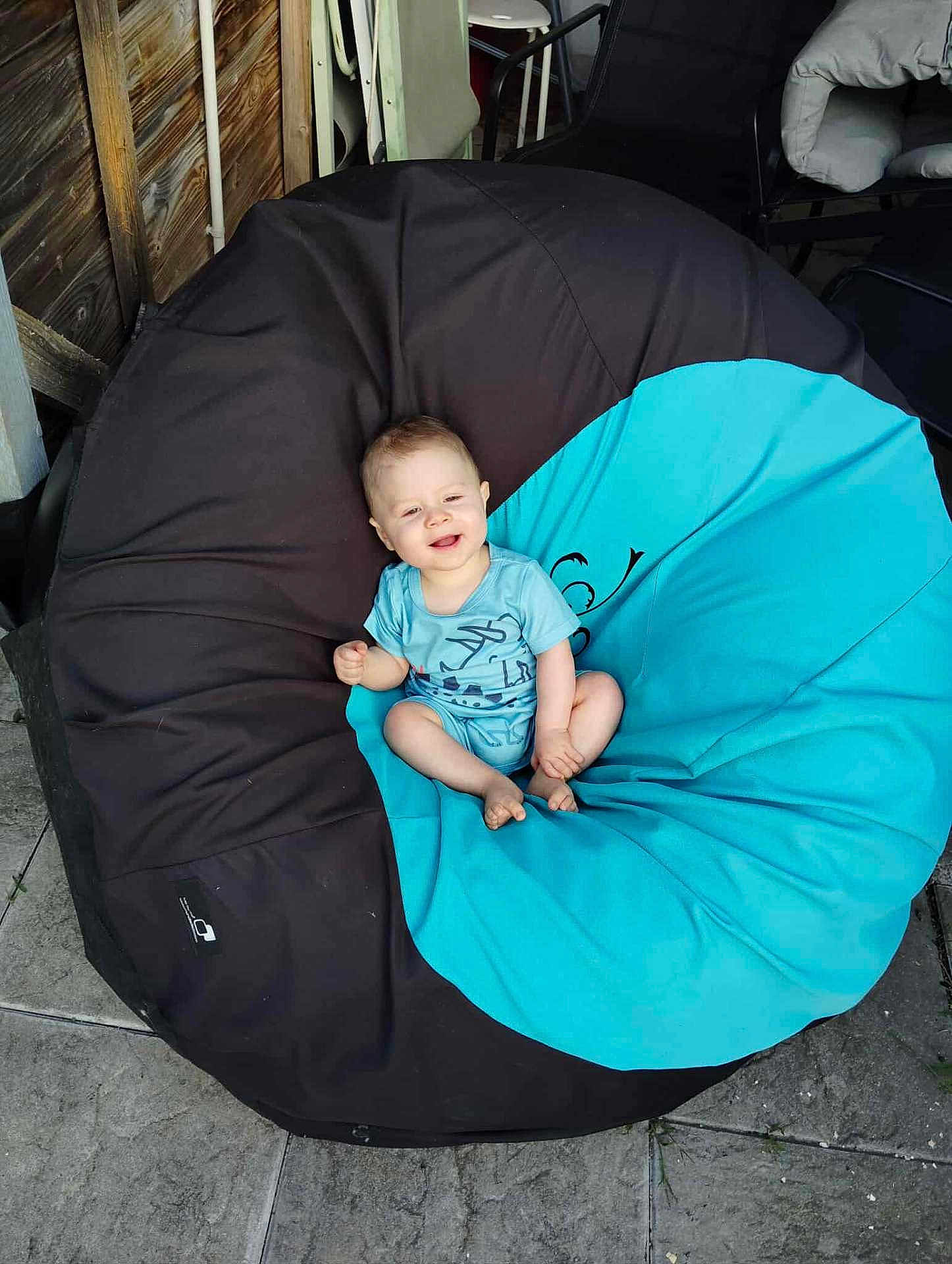 Gabriel a rejoint le concours — aidez-le/la à gagner de superbes lots ! baby, child, bean_bag, blue_clothing, outdoor, happy, smiling, sitting, floor, stone_tiles, wooden_wall, chair, furniture, person, casual, relaxed, infant, comfort, seat, sunlight