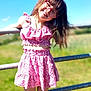 child, girl, smile, glasses, pink_clothing, dress, sunny, outdoor, fence, grass, blue_sky, happy, playful, summer, fashion, portrait, young, nature, casual, daylight