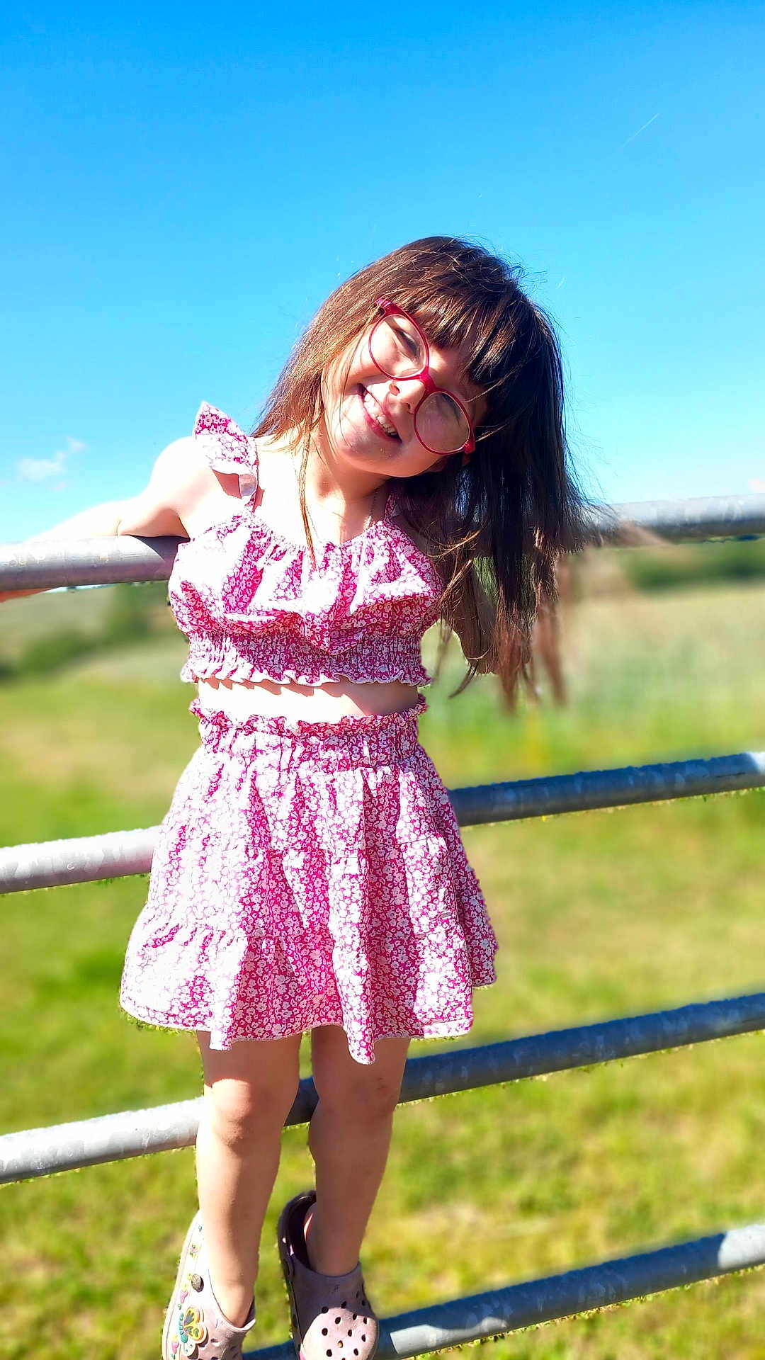 Lilia a rejoint le concours — aidez-le/la à gagner de superbes lots ! child, girl, smile, glasses, pink_clothing, dress, sunny, outdoor, fence, grass, blue_sky, happy, playful, summer, fashion, portrait, young, nature, casual, daylight