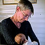 elderly_person, baby, indoor, portrait, affection, glasses, short_hair, black_sweater, table, painting, head, skin, person, family, love, resting, caring, seated, warmth, gentle