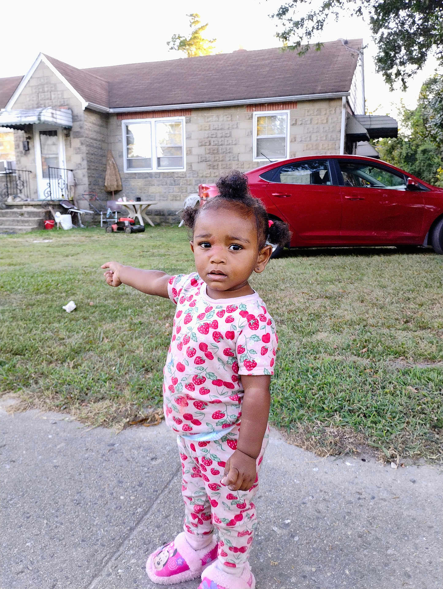 Miracle is registered to the contest to win money with this photo: toddler, child, pajamas, slippers, sidewalk, grass, house, car, pointing, outdoor, daylight, curly_hair, expression, clothing, footwear, person, yard, residential, suburban, casual