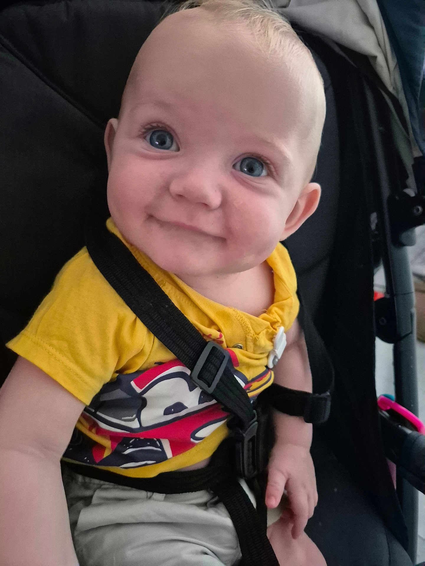Henry is registered to the contest to win money with this photo: baby, child, smiling, blue_eyes, yellow_shirt, stroller, seatbelt, cute, infant, happy, portrait, closeup, person, sitting, casual_clothing, indoors, young_child, face, hand, playful