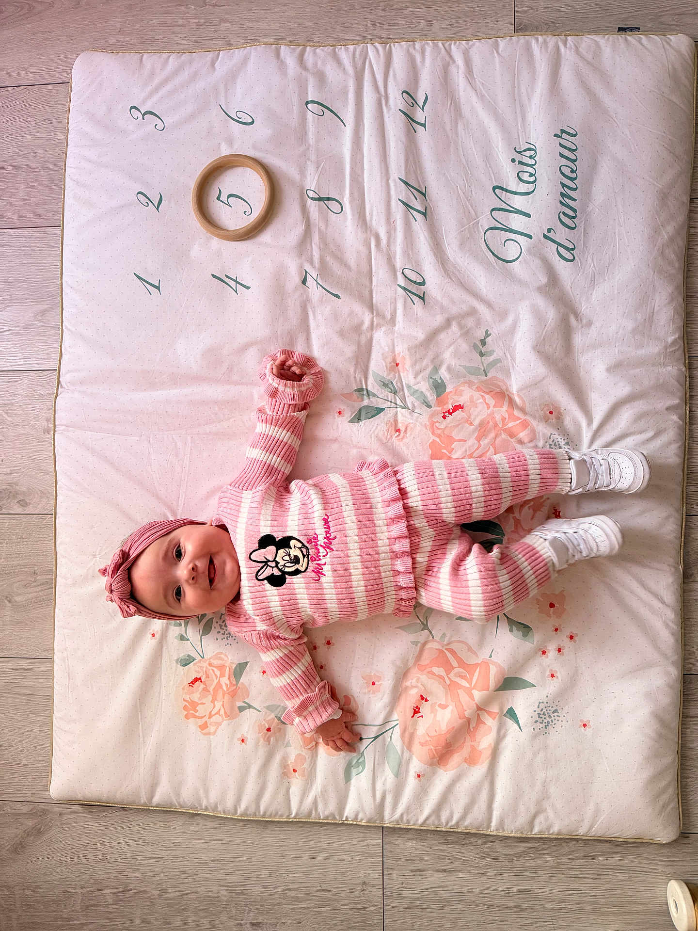 Gina participe au concours pour gagner de l'argent avec cette photo : baby, child, pink_clothing, striped_outfit, headband, smiling, lying_down, playmat, floral_pattern, numbers, wooden_floor, white_shoes, cute, infant, person, indoor, soft_mat, toy_ring, happy, portrait