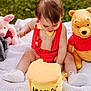 baby, child, cake, birthday, outdoor, grass, blanket, plush_toy, winnie_the_pooh, piglet_toy, red_suspender, yellow_bow_tie, white_shoes, cute, celebration, food, smash_cake, playful, sitting, curious