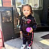 child, city, colorful, curly_hair, doorway, fashion, jacket, outdoor, portrait, reflection, smiling, sneakers, standing, steps, storefront, street, sunlight, toddler, toy, tracksuit