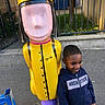 child, boy, hoodie, smile, outdoor, playground, statue, colorful, yellow, purple, sidewalk, fence, casual_clothing, person, urban, daylight, happy, standing, toy_figure, metal
