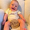 baby, infant, three_months, smiling, onesie, wooden_sign, milestone, sitting, plush_chair, cozy, cute, happy, child, portrait, indoor, soft_texture, chubby_cheeks, bright_eyes, light_clothing, closeup