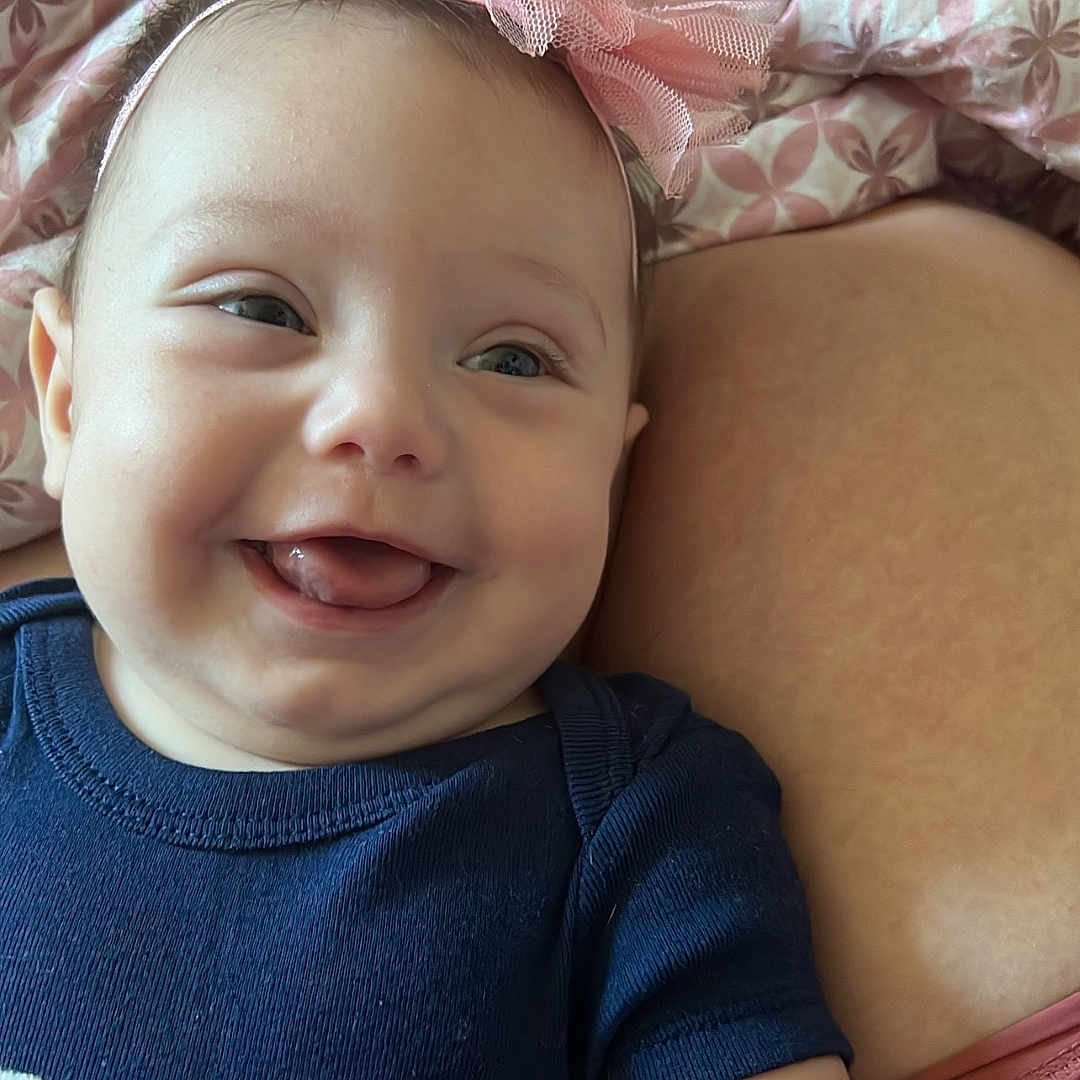 Zurriah is registered to the contest to win money with this photo: baby, blanket, cheek, closeup, cute, face, happy, headband, human, indoors, infant, lying_down, navy_blue_clothing, onesie, patterned_blanket, pink_bow, skin, smiling, soft_toy, tongue_out