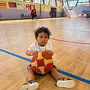 Ilyan participe au concours pour gagner de l'argent avec cette photo : toddler, child, basketball, gym, sports, court, indoor, shoes, curly_hair, white_shirt, blue_shorts, sandals, wooden_floor, windows, natural_light, basketball_hoop, kids, people, walking, sportswear
