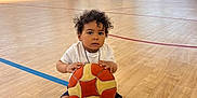 Ilyan a rejoint le concours — aidez-le/la à gagner de superbes lots ! toddler, child, basketball, gym, sports, court, indoor, shoes, curly_hair, white_shirt, blue_shorts, sandals, wooden_floor, windows, natural_light, basketball_hoop, kids, people, walking, sportswear