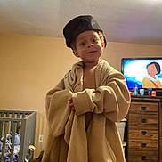 Lyric is registered to the contest to win money with this photo: child, indoor, oversized_clothing, jacket, cap, television, cartoon, crib, dresser, room, furniture, playful, person, face, headwear, brown_hair, casual, young, smile, home