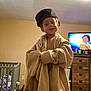 child, indoor, oversized_clothing, jacket, cap, television, cartoon, crib, dresser, room, furniture, playful, person, face, headwear, brown_hair, casual, young, smile, home