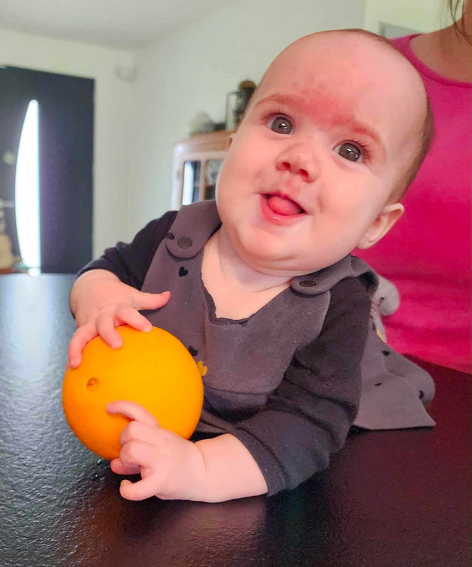 Joséphine participe au concours pour gagner de l'argent avec cette photo : baby, infant, orange_fruit, tongue_out, smiling, black_clothing, indoor, table, person, pink_clothing, face, hand, cute, playful, skin, head, eyes, expression, background, furniture
