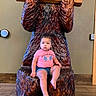 baby, child, infant, sitting, portrait, face, curious, indoor, wooden_throne, carved_wood, bear_sculpture, lodge, sign, pink_shirt, denim_shorts, barefoot, wall, floor, furniture, photograph