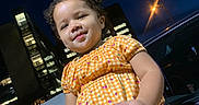 Damariana is registered to the contest to win money with this photo: child, toddler, girl, portrait, smiling, outdoor, night, city, street, car, dress, orange_clothing, checkered_pattern, curly_hair, standing, face, person, streetlight, buildings, urban_background