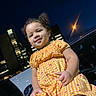 child, toddler, girl, portrait, smiling, outdoor, night, city, street, car, dress, orange_clothing, checkered_pattern, curly_hair, standing, face, person, streetlight, buildings, urban_background