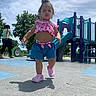 child, toddler, playground, outdoor, park, walking, standing, smiling, curly_hair, pink_clothing, shorts, sneakers, slide, playground_equipment, trees, blue_sky, sunny, shadow, portrait, daylight