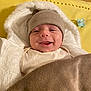baby, smile, hat, blanket, pacifier, yellow_mat, white_jacket, cozy, infant, child, warm_clothing, indoor, cute, face, headwear, wrapped, happy, lying_down, soft_texture, comfort