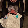 newborn, baby, infant, sleeping, car_seat, blanket, bow, headband, pacifier, portable_fan, parent_hand, indoor, portrait, face, eyes_closed, bundled, clothing, safety_straps, wood_floor, cozy