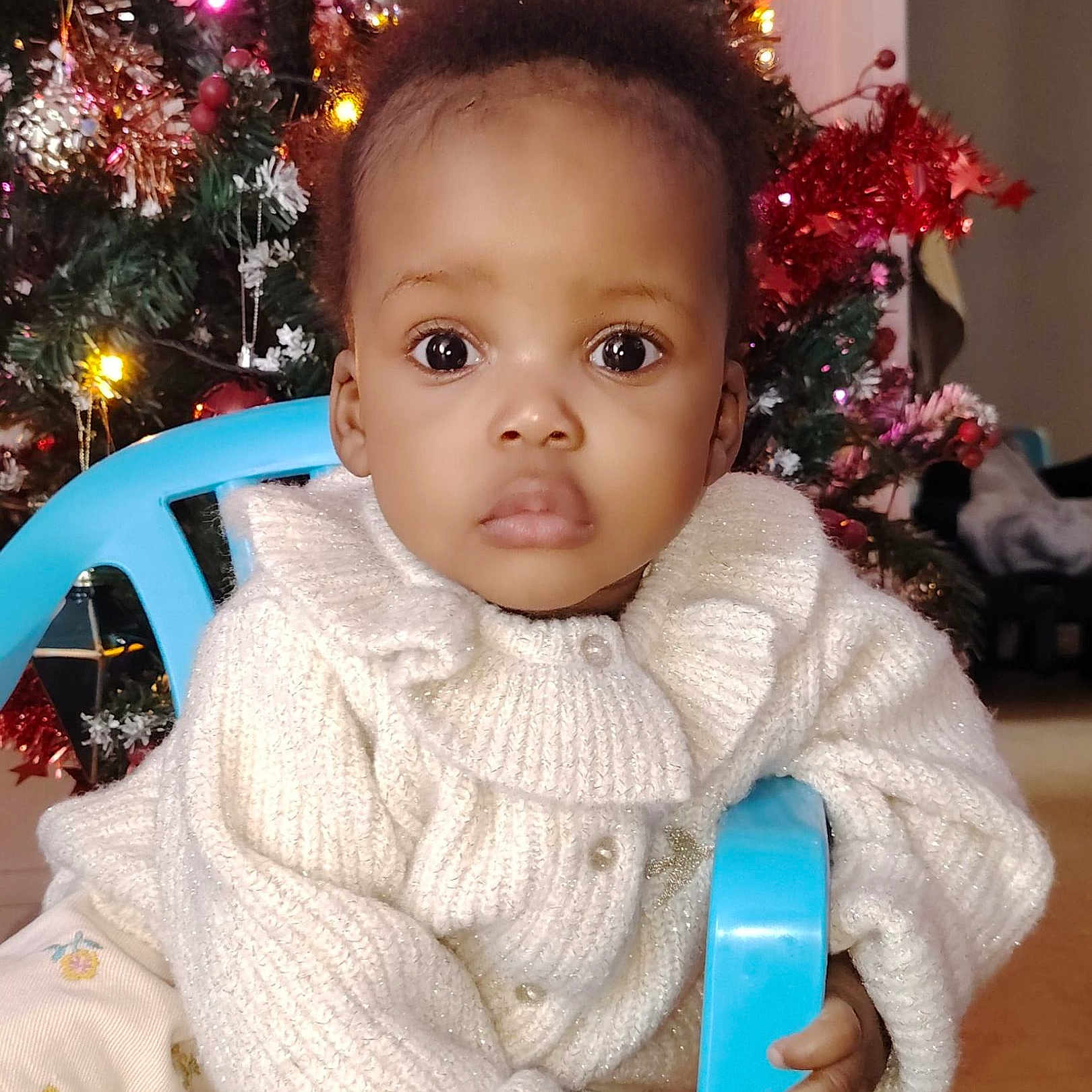 Syrielle a rejoint le concours — aidez-le/la à gagner de superbes lots ! baby, chair, child, christmas_tree, curly_hair, decorations, expression, eyes, face, festive, hand, holiday, home, indoors, ornaments, portrait, seated, sweater, toddler, white_clothing