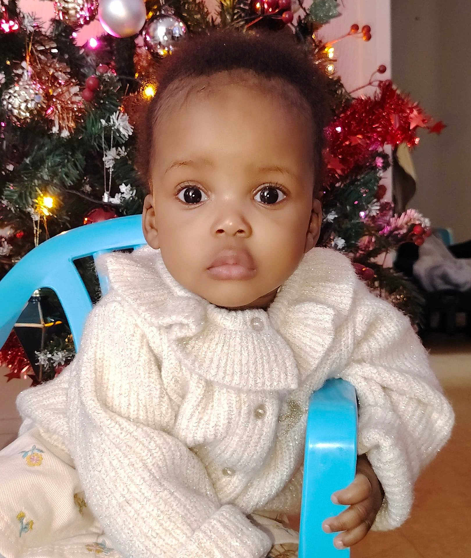 Syrielle a rejoint le concours — aidez-le/la à gagner de superbes lots ! toddler, child, baby, face, eyes, sweater, white_clothing, chair, hand, indoors, christmas_tree, ornaments, holiday, decorations, festive, curly_hair, expression, portrait, seated, home