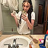 woman, cat, mirror, bathroom, selfie, sink, counter, toiletries, shower_curtain, tshirt, long_hair, phone, glasses, pet, reflection, door, hairbrush, basket, hand, neutral_expression