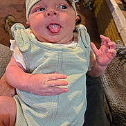 Nirvana is registered to the contest to win money with this photo: baby, infant, child, tongue_out, headband, bow, clothing, sleeveless, hand, face, skin, indoors, carpet, furniture, person, cute, playful, expression, holding, seated