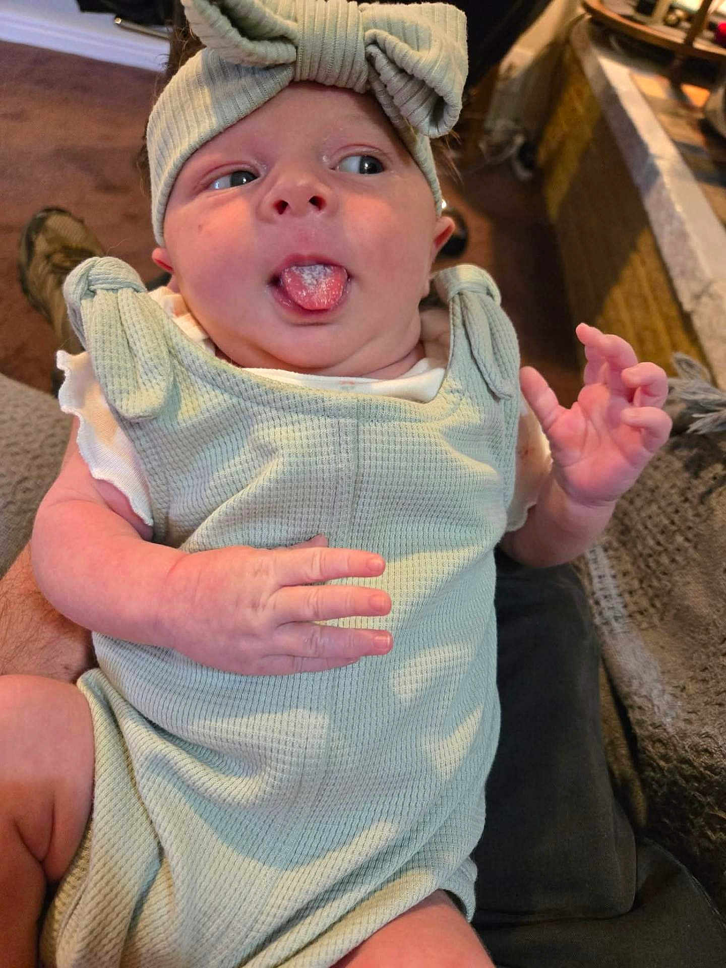 Nirvana is registered to the contest to win money with this photo: baby, infant, child, tongue_out, headband, bow, clothing, sleeveless, hand, face, skin, indoors, carpet, furniture, person, cute, playful, expression, holding, seated