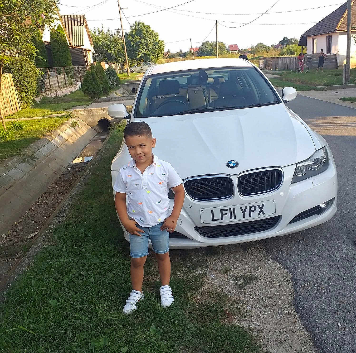 Zsolt is registered to the contest to win money with this photo: child, boy, car, bmw, white_car, street, grass, sidewalk, house, tree, daylight, casual_clothing, shorts, sneakers, smiling, outdoor, portrait, road, fence, urban