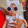 baby, child, sunglasses, headband, bow, hello_kitty, onesie, tattoo, hand, person, indoor, clothing, cute, fashion, accessory, infant, holding, portrait, face, funny