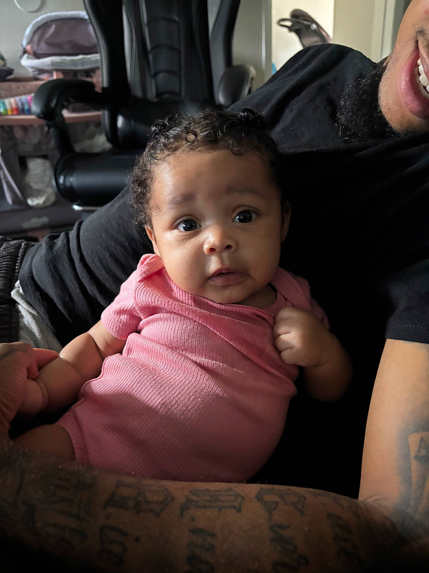 Kaziyah joined the competition — help win amazing prizes! adult, arm, baby, baby_gear, chair, closeup, clothing, curly_hair, cute, expression, holding, indoor, infant, person, pink_onesie, portrait, seated, skin, smile, tattoo