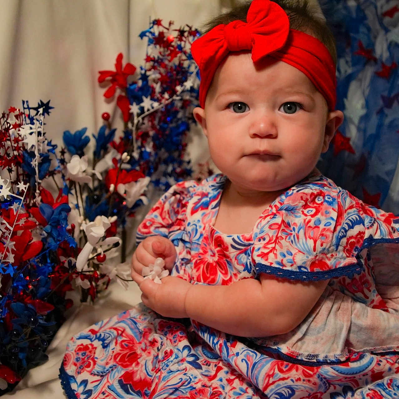 Emily is registered to the contest to win money with this photo: baby, child, infant, red_headband, floral_dress, blue, red, white, stars, decorations, patriotic, celebration, portrait, cute, sitting, face, hands, indoor, festive, holiday