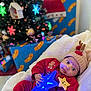 baby, child, christmas_tree, holiday, gift, present, red_clothing, reindeer_hat, star_wand, glowing_toy, blanket, indoor, festive, cute, smiling_star, decorations, ornament, christmas, holiday_lighting, cozy