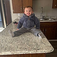 Hunter is registered to the contest to win money with this photo: toddler, child, smiling, kitchen, countertop, gray_clothing, fleece_jacket, fleece_pants, socks, wood_cabinets, sink, indoor, happy, person, portrait, home, apartment, flooring, door, lighting