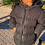 child, braided_hair, black_jacket, outdoor, park, sunlight, grass, pavement, person, thoughtful, casual_clothing, young, standing, cold_weather, nature, daylight, expression, face, portrait, winter