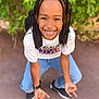 child, smiling, braided_hair, crouching, outdoor, greenery, casual_clothing, white_tshirt, blue_jeans, black_shoes, watch, black_backpack, happy, person, portrait, daylight, playful, young, friendly, urban