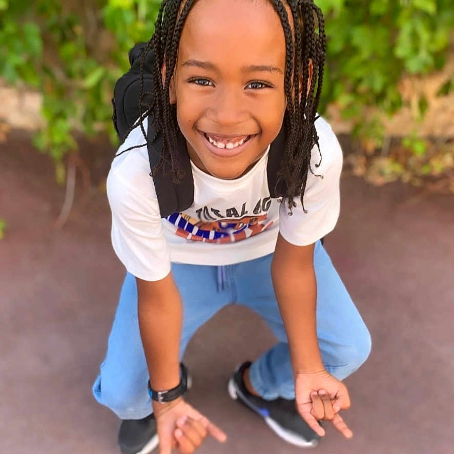 Jahïden a rejoint le concours — aidez-le/la à gagner de superbes lots ! black_backpack, black_shoes, blue_jeans, braided_hair, casual_clothing, child, crouching, daylight, friendly, greenery, happy, outdoor, person, playful, portrait, smiling, urban, watch, white_tshirt, young